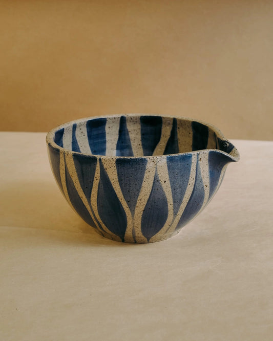 A handmade ceramic bowl with a blue and beige wavy vertical pattern, featuring a small pouring spout on the right side, set against a warm beige background.