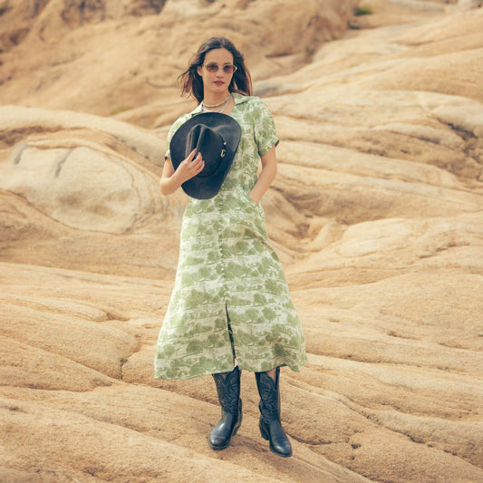 Woman in a green dress holding a black hat against a desert landscape, Woman wearing a green patterned dress and black cowboy hat and the Darcy Dress with feminine charm and a hint of retro style. Stylish dress cowboy rancher girl