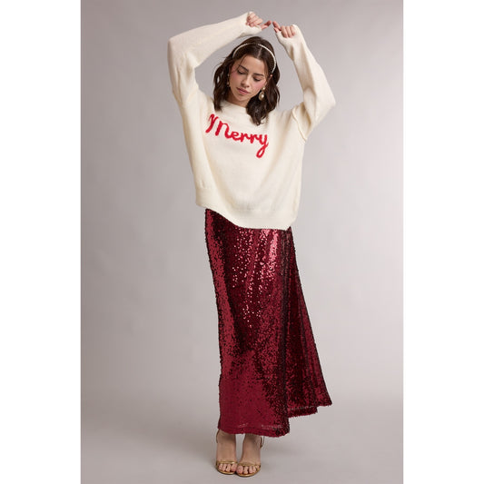 Woman with hands above her head wearing long sequin skirt with an of-white, holiday sweater with the word "Merry" written across it