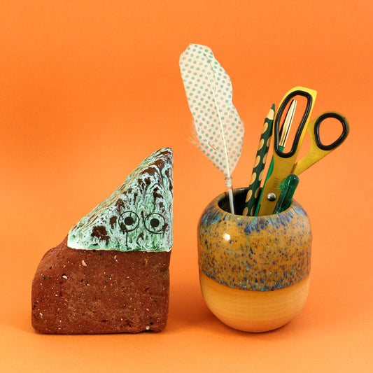 Ceramic rock with eyes and a ceramic cup with stationery items on an orange background
