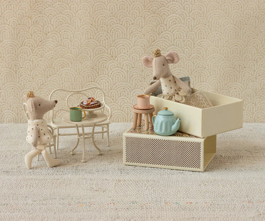 Miniature mouse figures with a tea set on a textured surface with a patterned wall background.