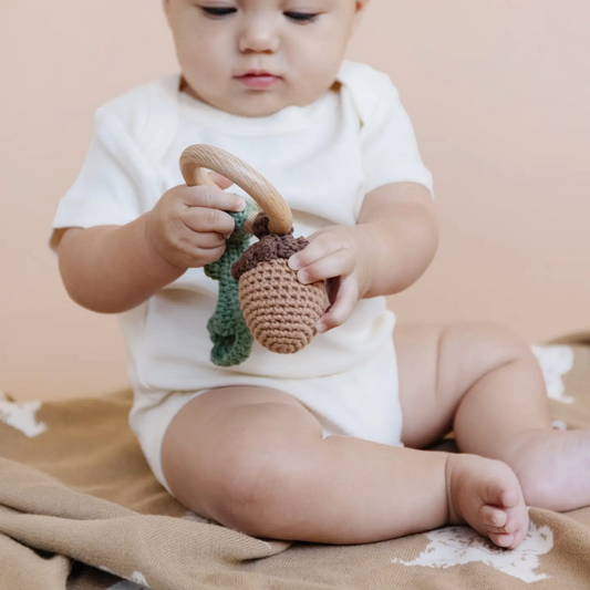 Baby playing with a wooden rattle and crocheted toy on a beige blanket. Showing off the tactile feel of the Blueberry Hill rattle and the cute acorn and leaf.