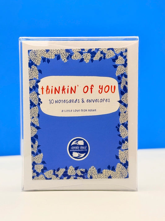 A boxed card set labeled "thinkin' of you" with 10 notecards and envelopes, shown upright against a bright blue background. The package has a blue front design with a white floral border and red and black text.