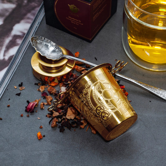 Gold tea cup with intricate designs on a dark surface with tea leaves and a glass of tea.