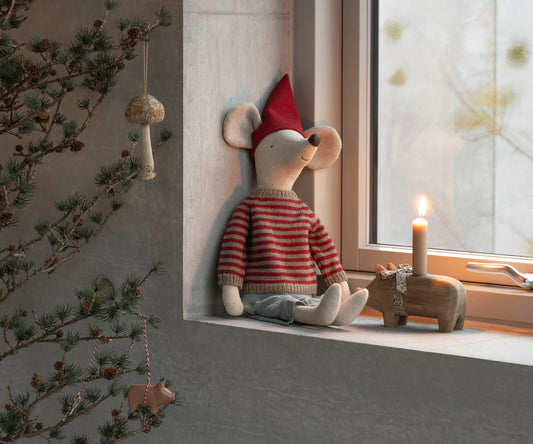 Stuffed mouse toy with a red hat and striped sweater sitting by a window with a candle and wooden toys.