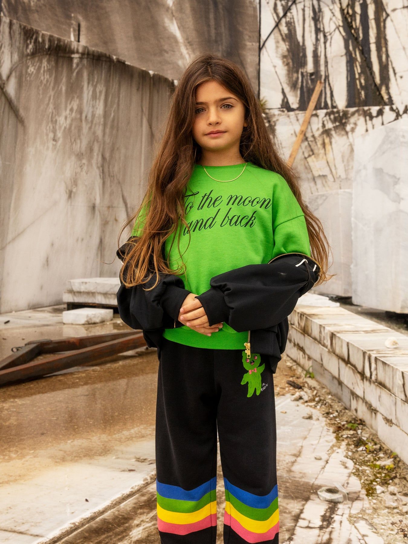 Child wearing a green sweatshirt with text and black pants with colorful stripes, standing in an outdoor setting.