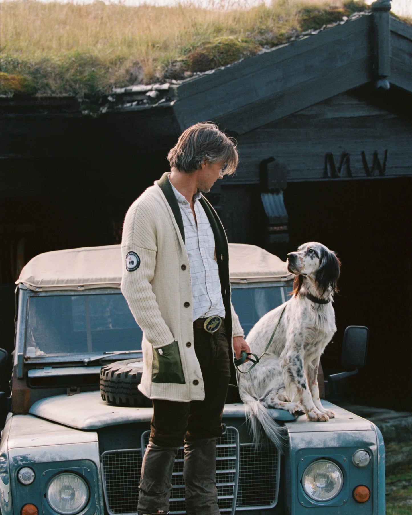 Man in a the new norwegian brand sweater and cardigan standing next to a dog on an old Land Rover in a rustic setting. The cozy nature of this sweater is perfect for fall 2025 and men's fashion