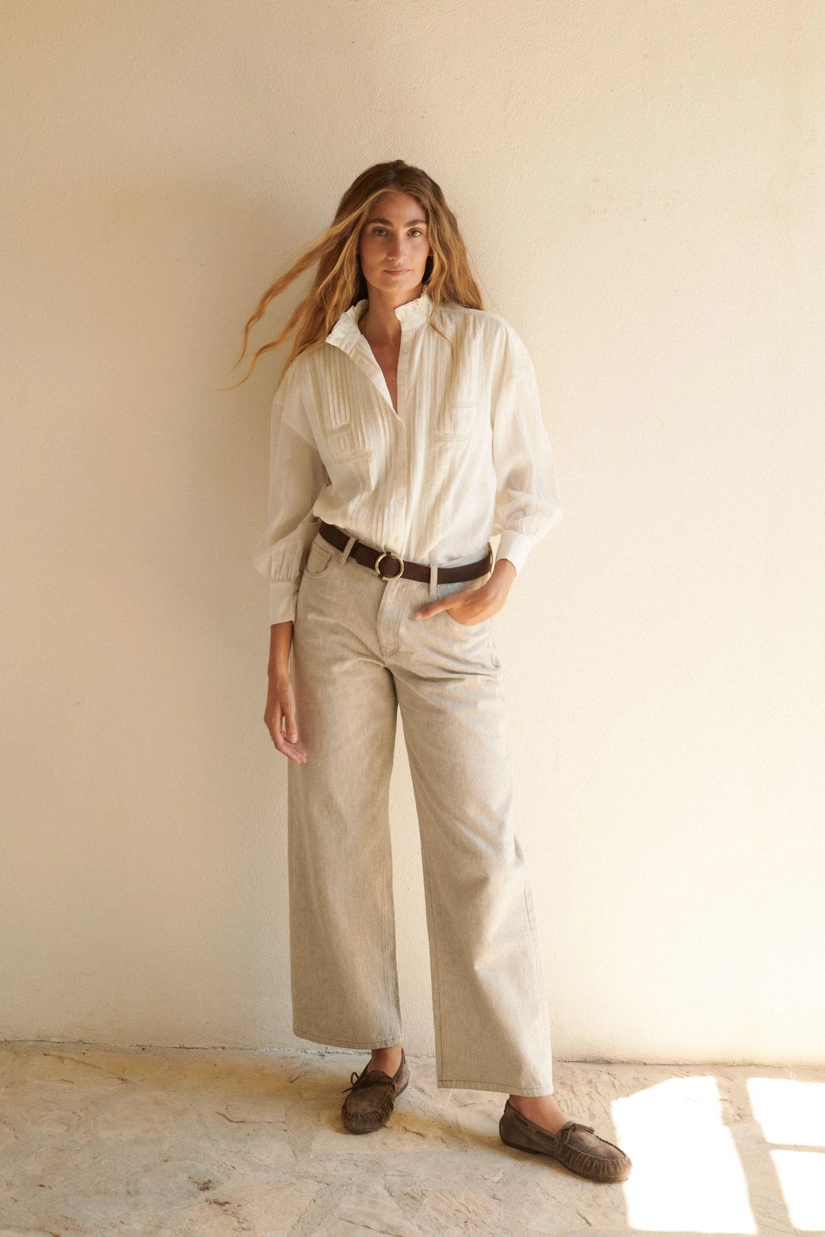 Woman wearing a white shirt and beige pants standing in a sunlit room. Alexa Blouse is bohemian and airy for spring and summer or hot weather.
