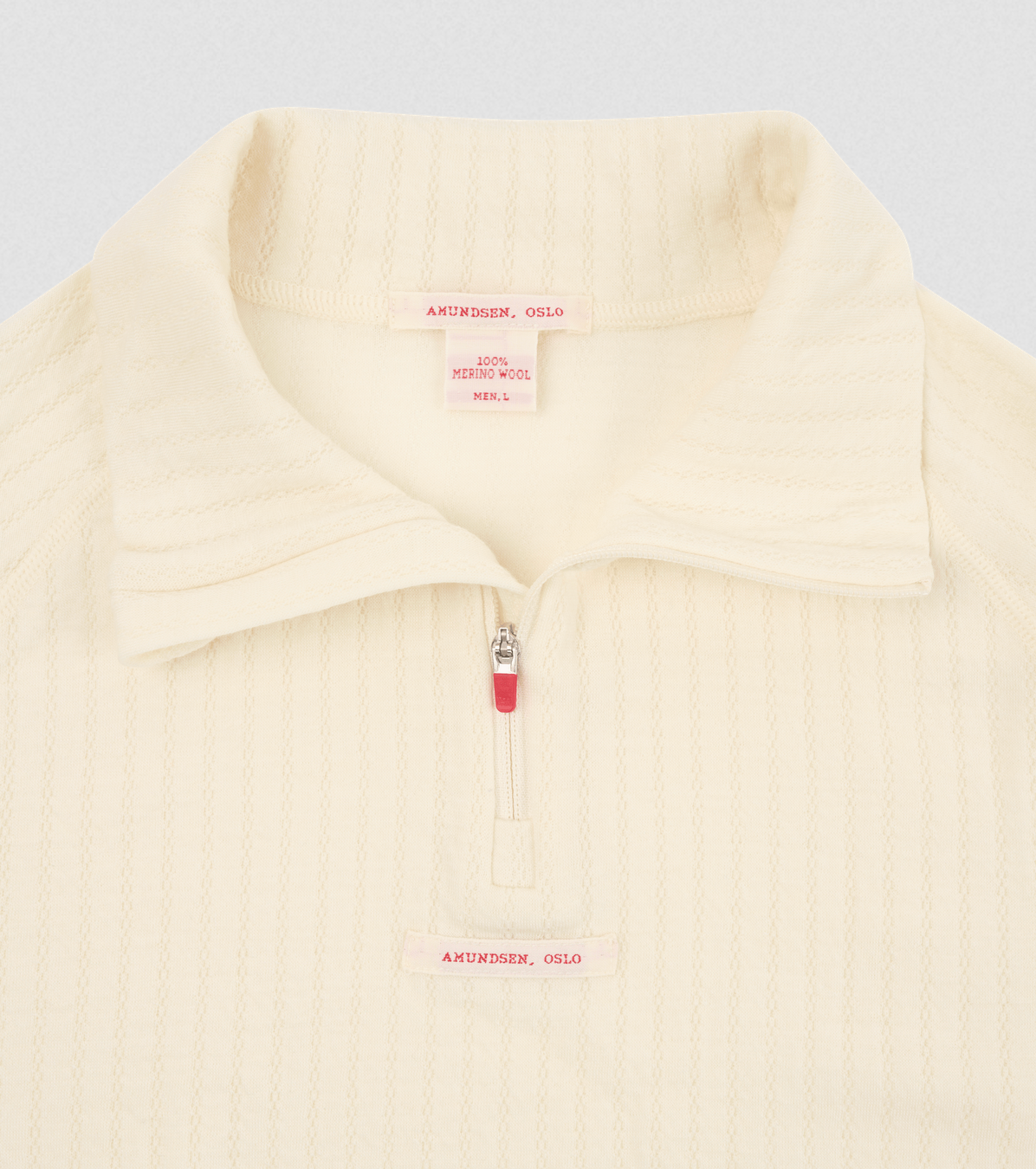 Close-up of a cream-colored ribbed knit quarter-zip pullover with a collar, featuring a small pink brand label and zipper pull on a light gray background.