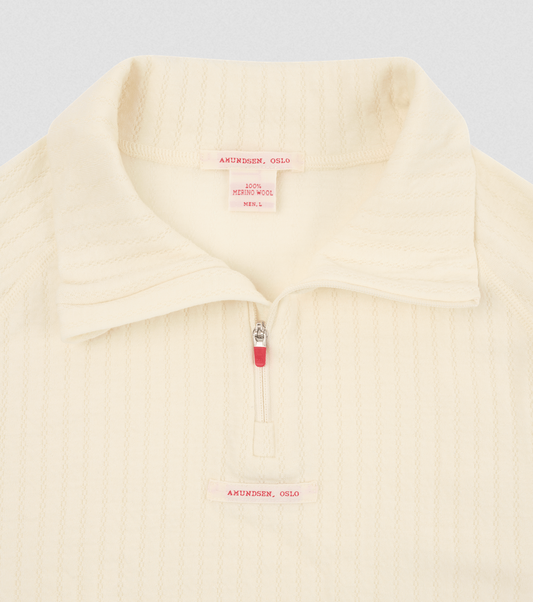 Close-up of a cream-colored ribbed knit quarter-zip pullover with a collar, featuring a small pink brand label and zipper pull on a light gray background.