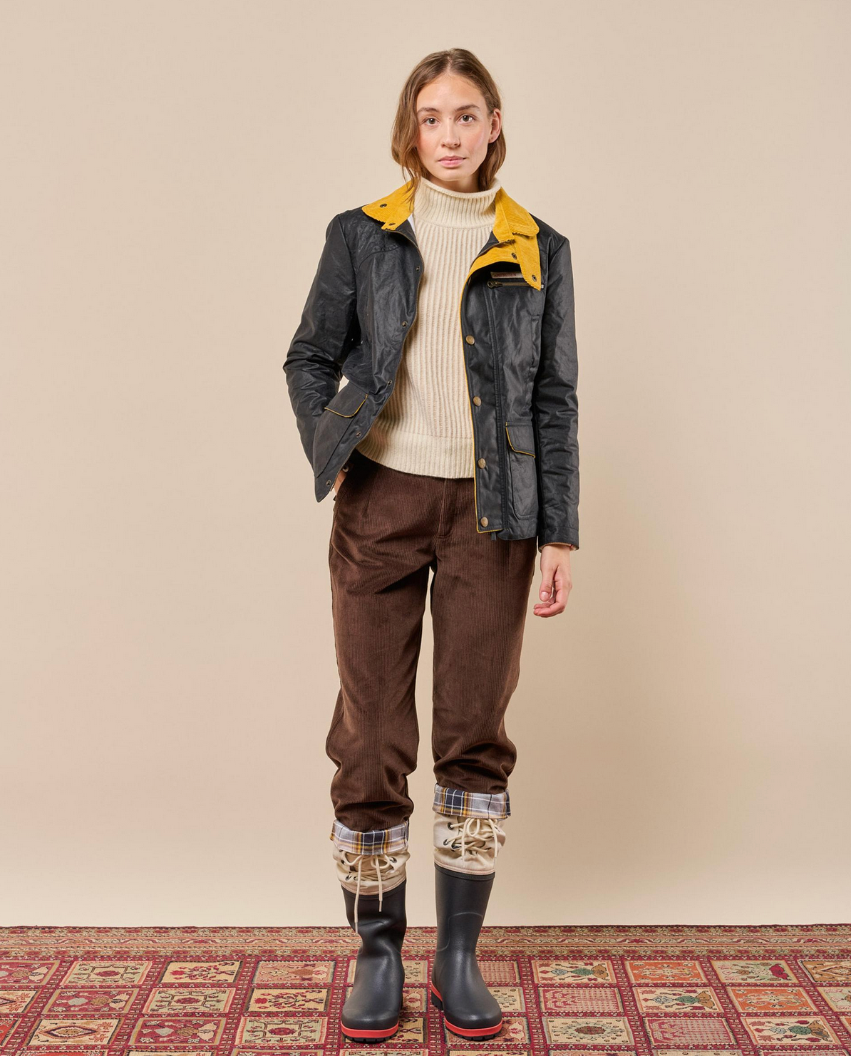 A model stands against a plain beige studio wall wearing a dark waxed jacket with a bright yellow collar, a cream ribbed turtleneck, brown corduroy pants, plaid cuff socks, and black rubber rain boots with red soles, standing on a patterned rug.