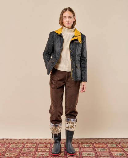 A model stands against a plain beige studio wall wearing a dark waxed jacket with a bright yellow collar, a cream ribbed turtleneck, brown corduroy pants, plaid cuff socks, and black rubber rain boots with red soles, standing on a patterned rug.