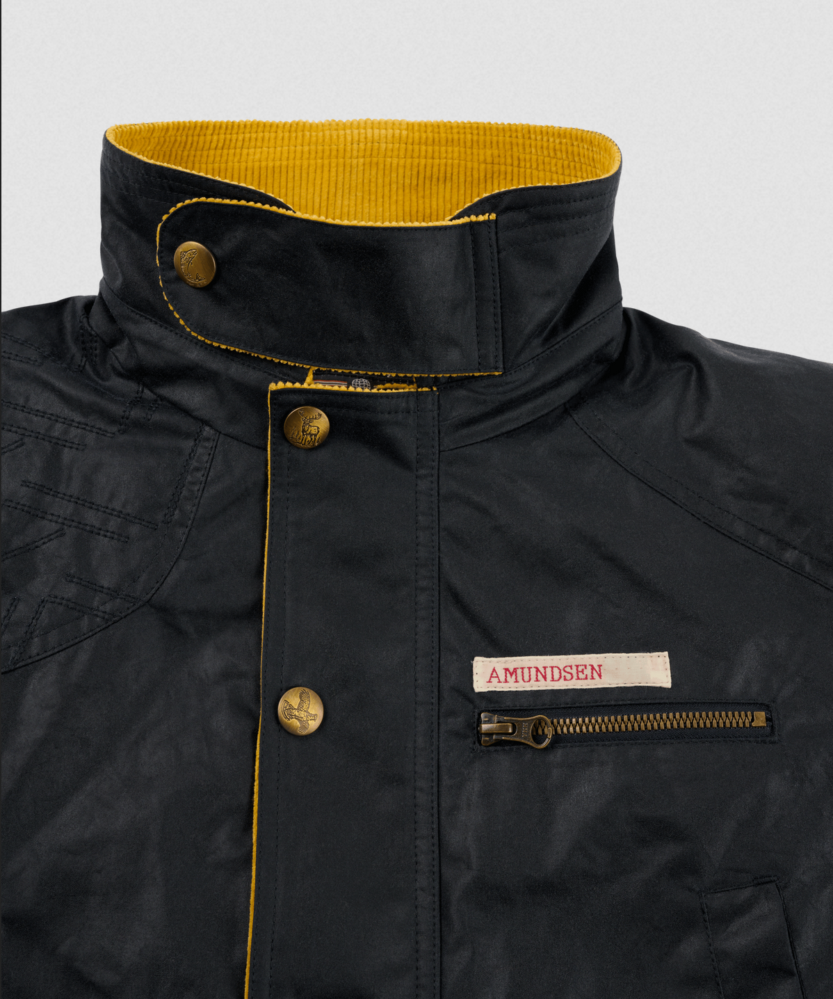 Close-up of a black waxed jacket with a high collar, mustard yellow corduroy lining, brass buttons, and a zippered chest pocket. A white label on the chest reads 'AMUNDSEN'.