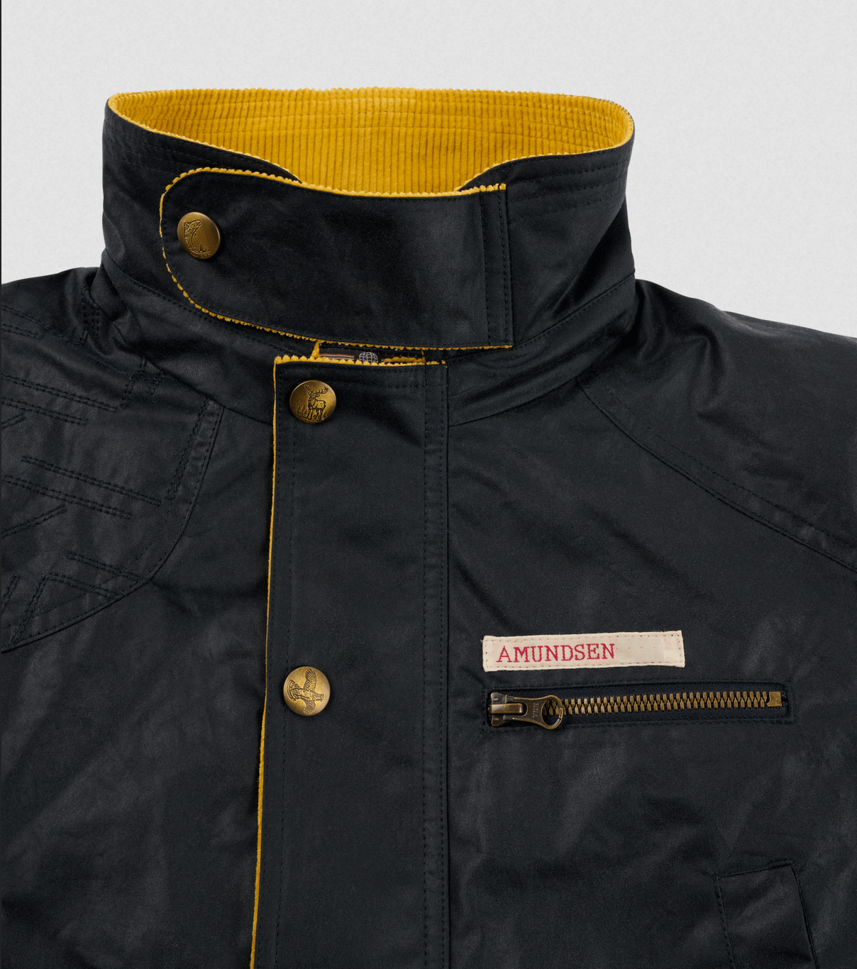 Close-up of a black waxed jacket with a high collar, yellow interior lining, brass buttons, and a zippered chest pocket featuring a white AMUNDSEN label.