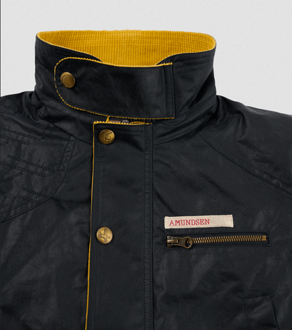 Close-up of a black waxed jacket with a high collar, yellow interior lining, brass buttons, and a zippered chest pocket featuring a white AMUNDSEN label.