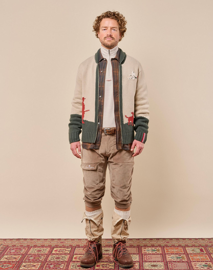 A man stands facing forward wearing a beige knit cardigan with dark green ribbed trim, a white mock-neck shirt, brown plaid trousers, and brown lace-up boots, posed against a plain beige studio background with a patterned rug at the bottom.