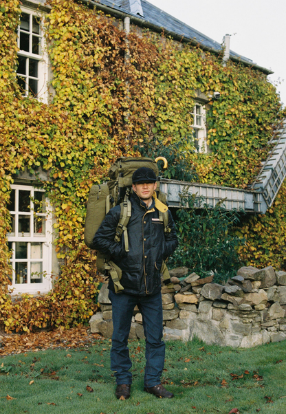 A man wearing a large olive green hiking backpack and a black jacket stands in front of a stone building covered in green and yellow ivy.