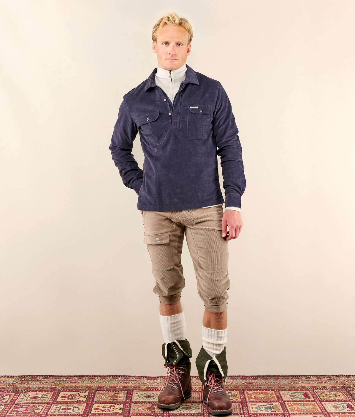 A man stands against a plain beige studio wall wearing a navy blue long-sleeve button-front overshirt over a white mock neck, tan cropped cargo pants, white socks, and dark green and brown boots. A patterned rug is visible at the bottom.