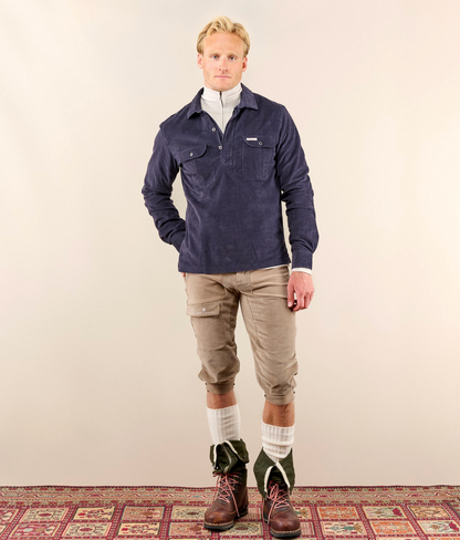 A man stands against a plain beige studio wall wearing a navy blue long-sleeve button-front overshirt over a white mock neck, tan cropped cargo pants, white socks, and dark green and brown boots. A patterned rug is visible at the bottom.