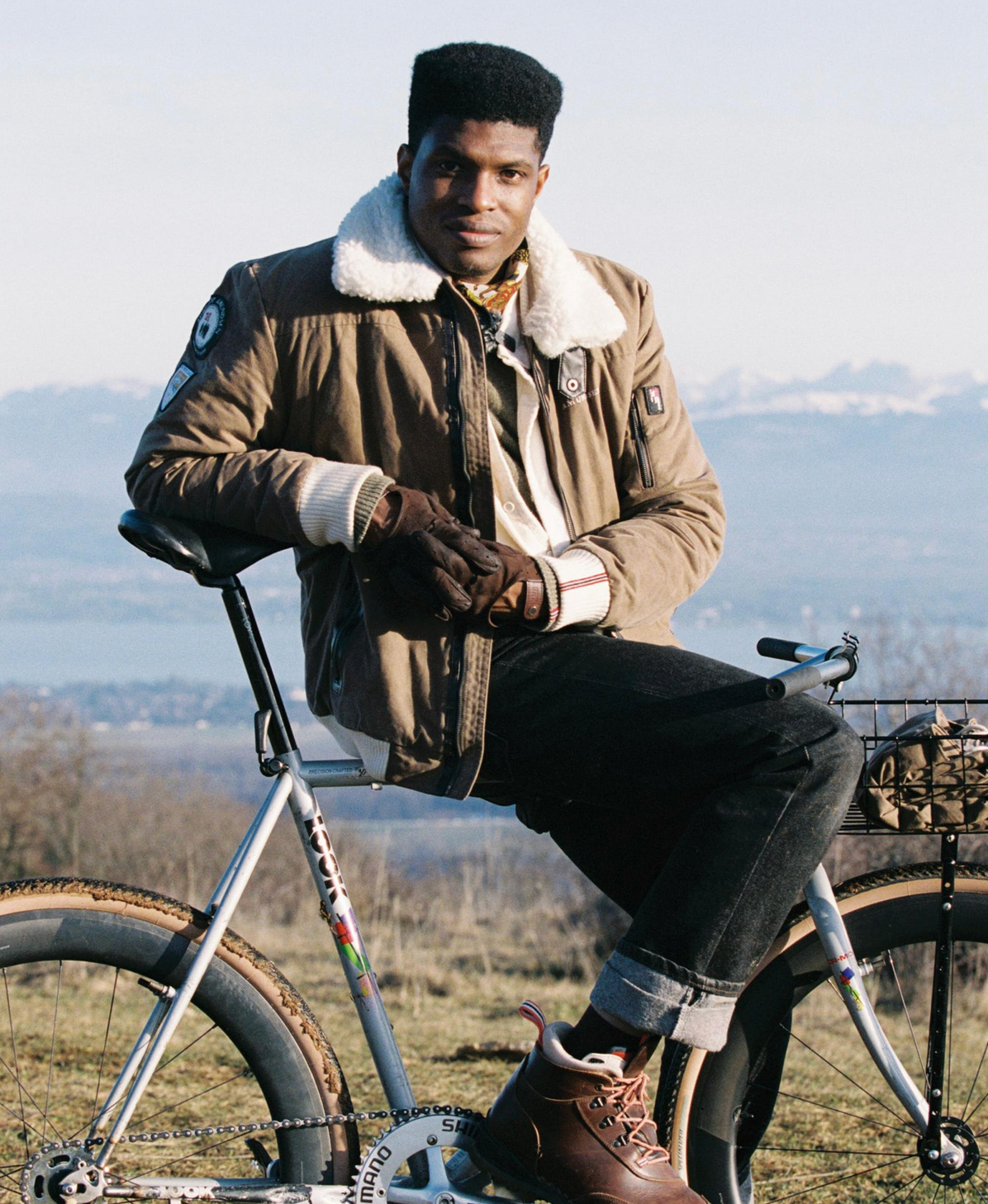 A man wearing a brown shearling-lined jacket, black jeans, and brown boots sits on a silver bicycle outdoors with a mountain landscape in the background.