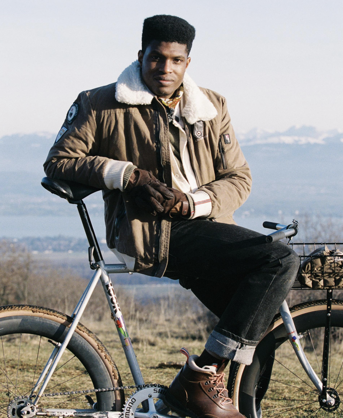 A man wearing a brown shearling-lined jacket, black jeans, and brown boots sits on a silver bicycle outdoors with a mountain landscape in the background.