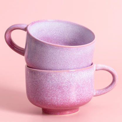 Two stacked ceramic mugs with a textured, speckled finish in shades of pink and purple, set against a solid pastel pink background.