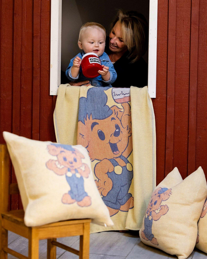 Woman holding a baby with a teddy bear-themed blanket and pillows in a room with wooden walls.