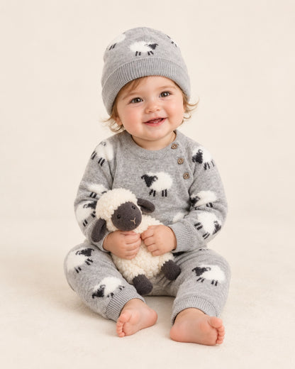 A baby sitting on a cream studio background wearing a gray knit one-piece pajama set with black and white sheep motifs, a matching gray beanie, and holding a small plush sheep toy.