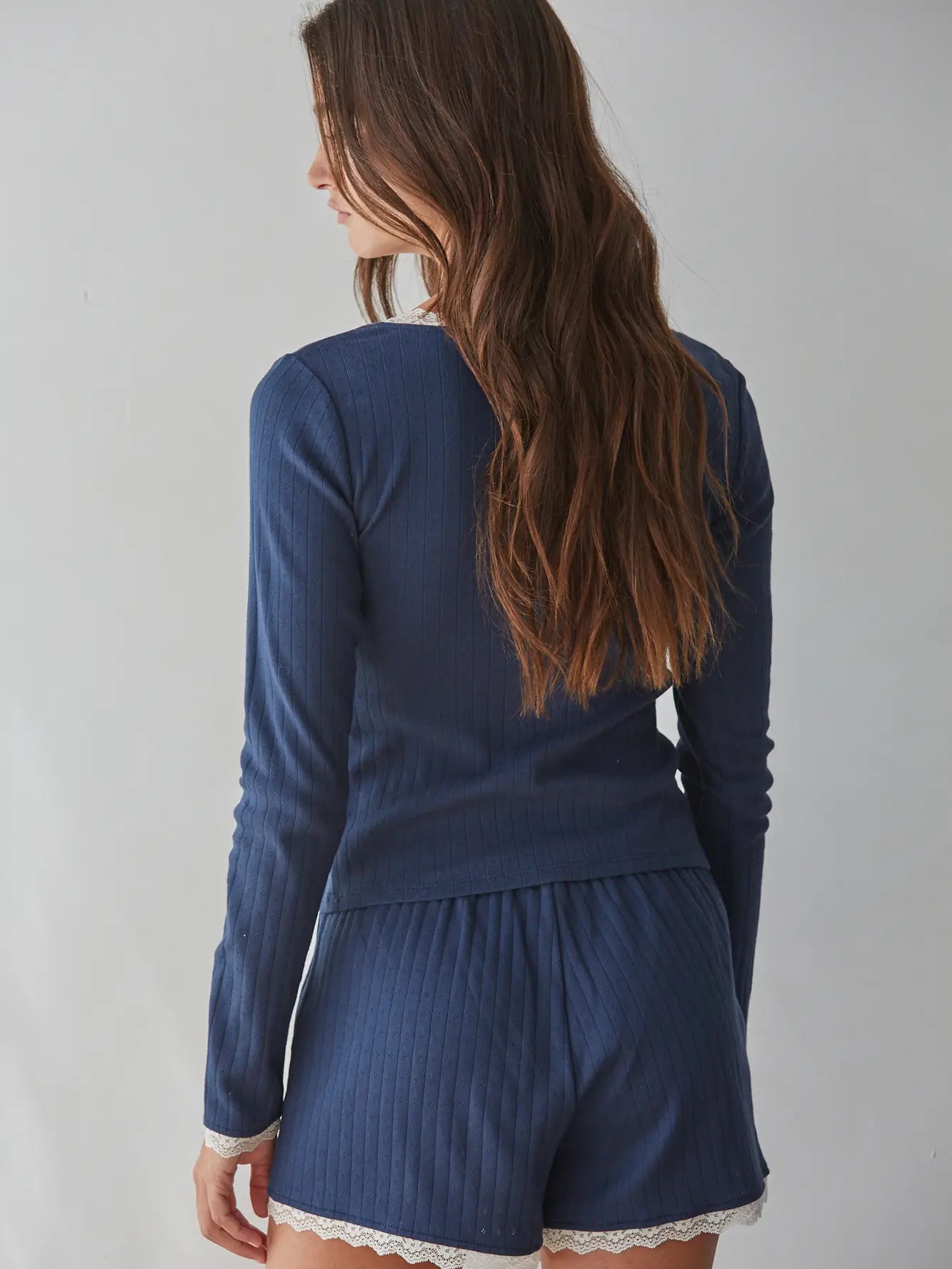 A woman shown from the back wearing a matching navy blue ribbed long-sleeve top and shorts set with white lace trim, against a plain light gray background.