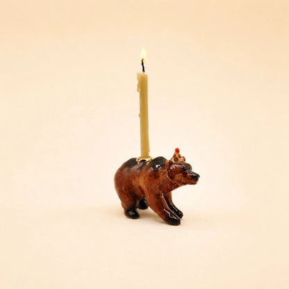 A bear-shaped candle holder with a lit cream-colored candle inserted into its back, set against a solid light beige background.