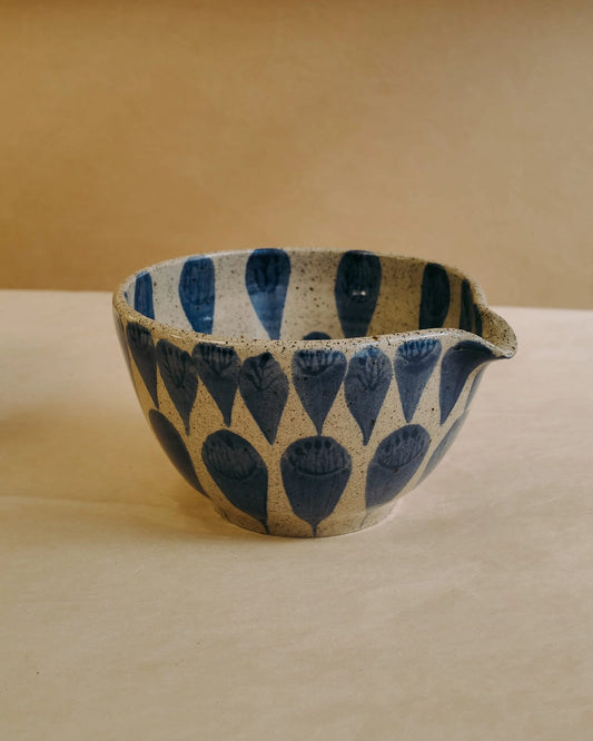 A handmade ceramic matcha bowl with a blue and beige speckled pattern, featuring a small pouring spout, set against a warm beige background.