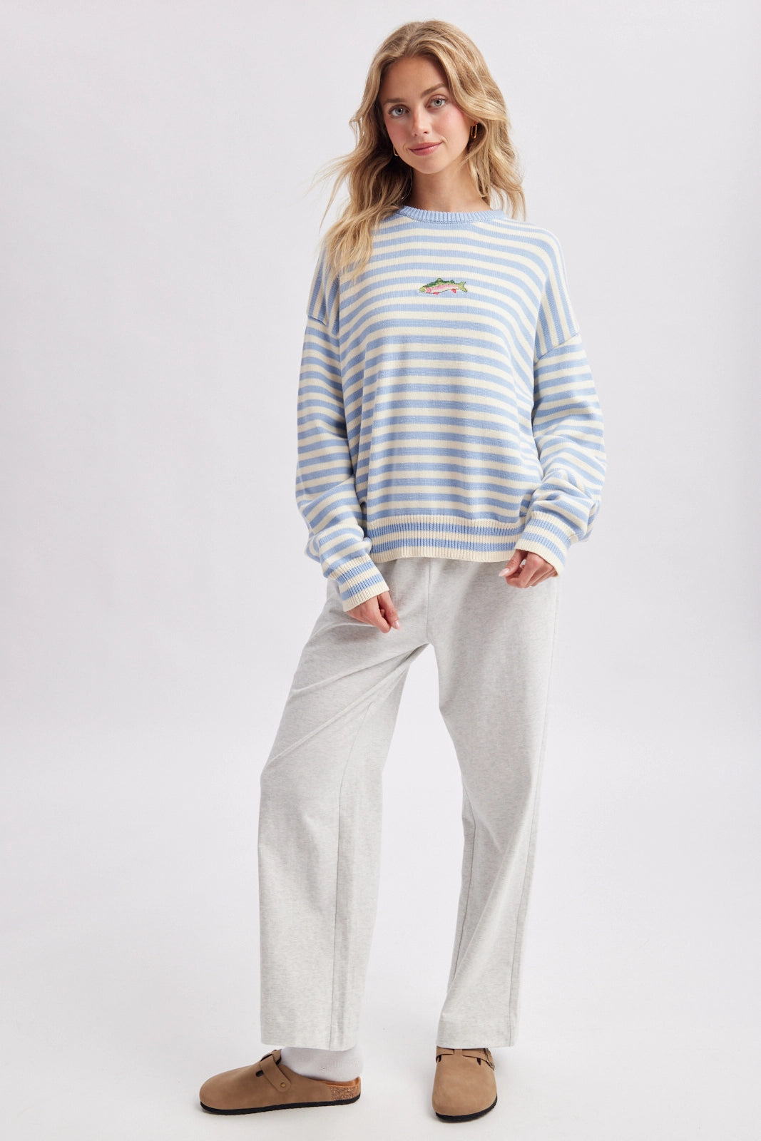 A woman modeling a light blue and white striped long-sleeve sweatshirt with a small embroidered logo on the left chest, paired with light gray sweatpants and brown mule shoes, against a plain light gray studio background.