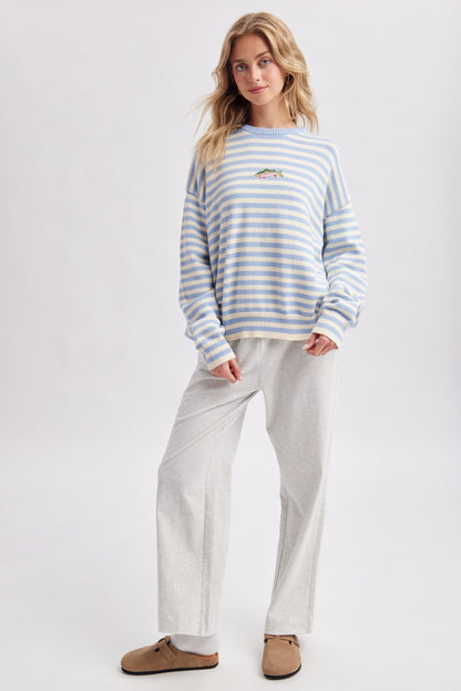 A woman modeling a light blue and white striped long-sleeve sweatshirt with a small embroidered logo on the left chest, paired with light gray sweatpants and brown mule shoes, against a plain light gray studio background.
