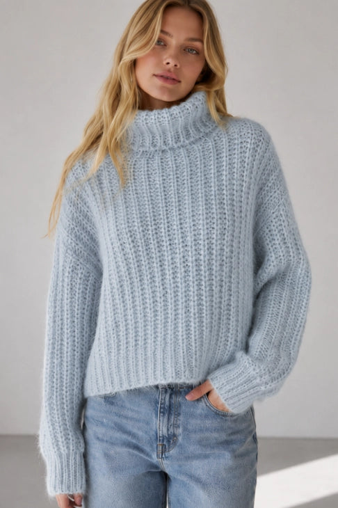 A woman wearing a light blue chunky knit mock neck sweater and light wash blue jeans, standing against a neutral light gray background.