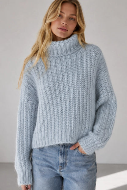 A woman wearing a light blue chunky knit mock neck sweater and light wash blue jeans, standing against a neutral light gray background.