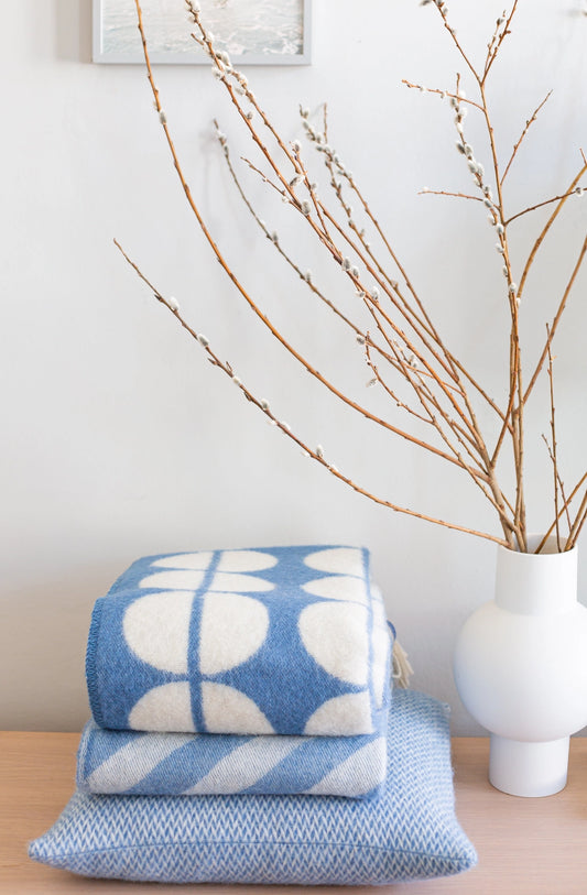 A stack of folded blue and white patterned textiles, including a throw blanket and a pillow, displayed on a light wood surface next to a white vase with dried branches against a white wall.
