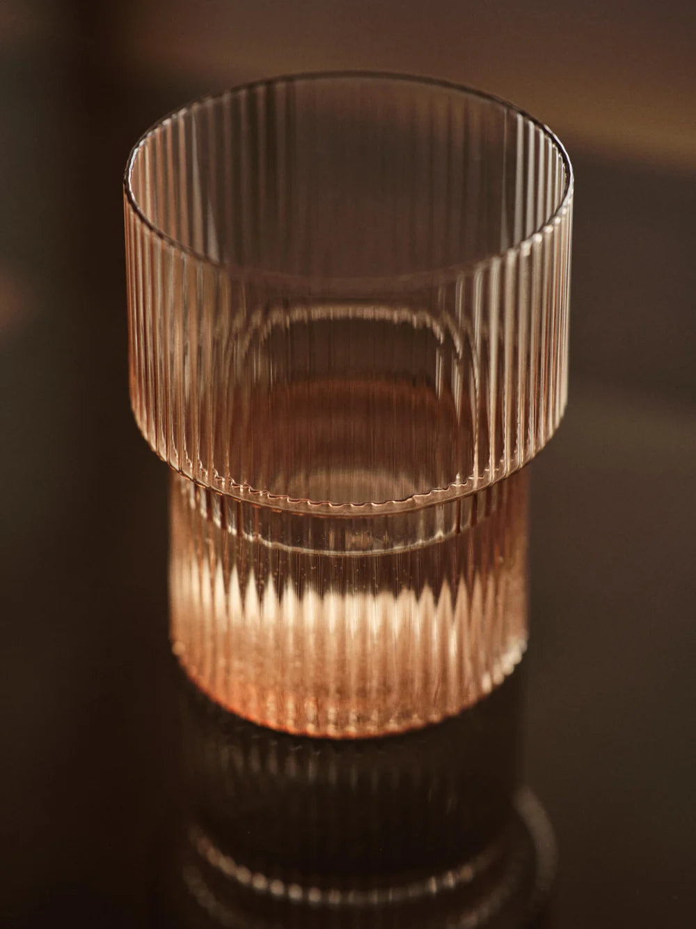 A clear ribbed glass tumbler with a warm amber tint, shown on a dark reflective surface in moody warm lighting.