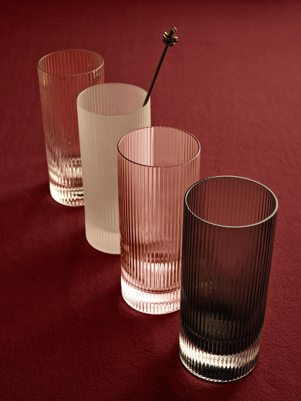 Four ribbed glass tumblers in different translucent colors arranged on a dark red background, with a metal stirrer resting in one of the glasses.