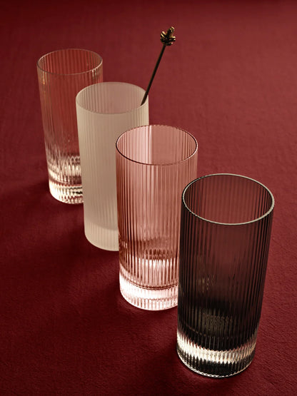 Four ribbed glass tumblers in different translucent colors arranged on a dark red background, with a metal stirrer resting in one of the glasses.