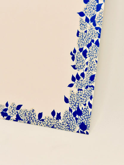 A close-up of a white square product corner decorated with a blue floral and leaf pattern, shown against a light beige background.