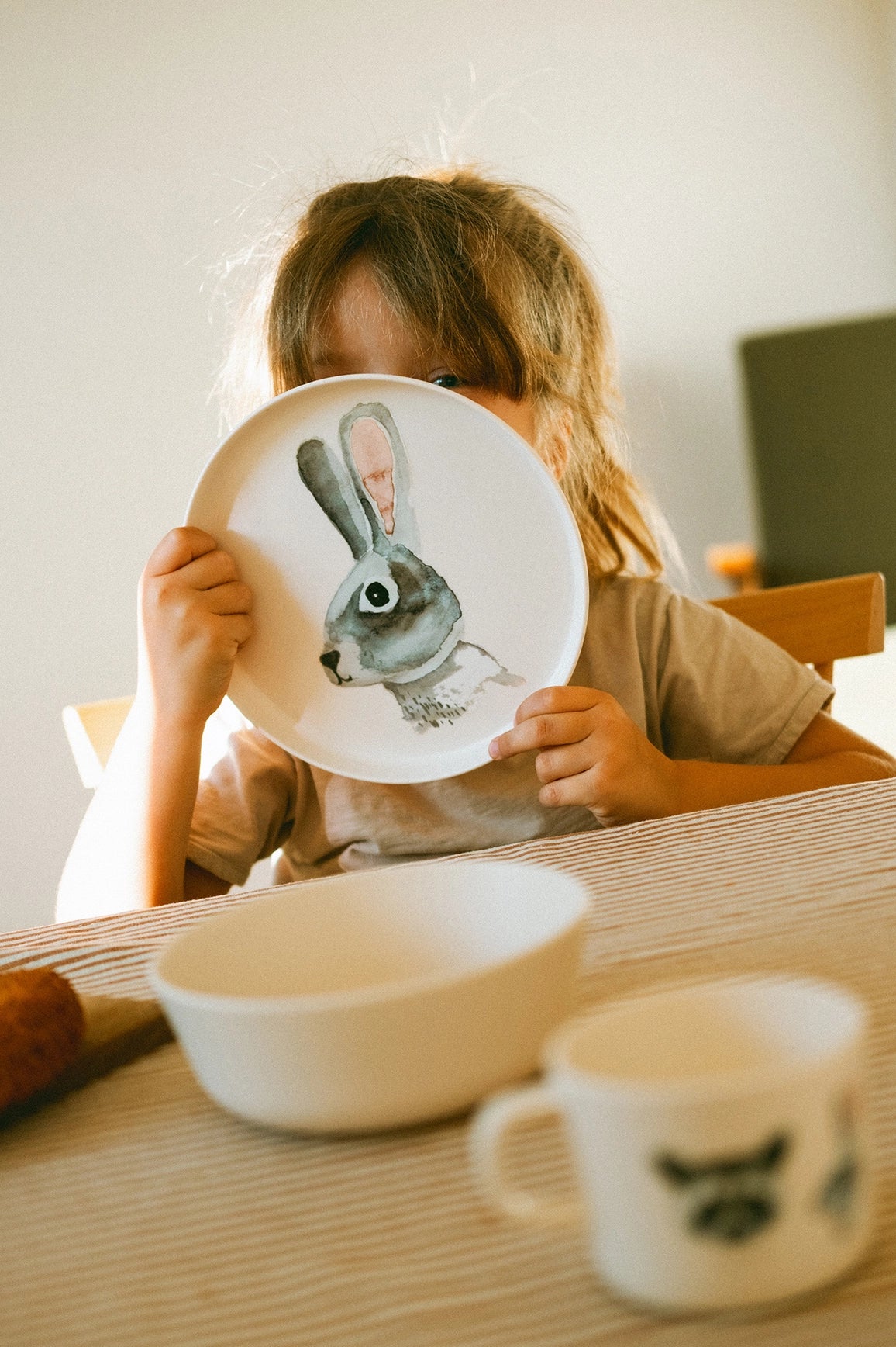 A child sits at a table holding a white plate with a painted gray rabbit in front of their face. A white bowl and a white mug with a dark graphic are on the table in the foreground.