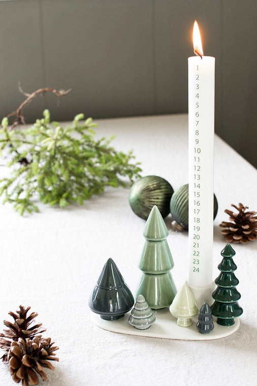 Decorative candle with number markers on a white surface with green ceramic trees and pinecones.
