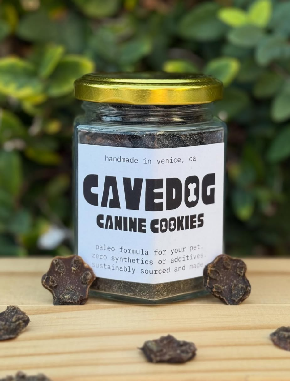 A glass jar of Cavedog Canine Cookies with a gold lid and white label, placed on a wooden surface with dog treats scattered around it and a blurred green garden background.