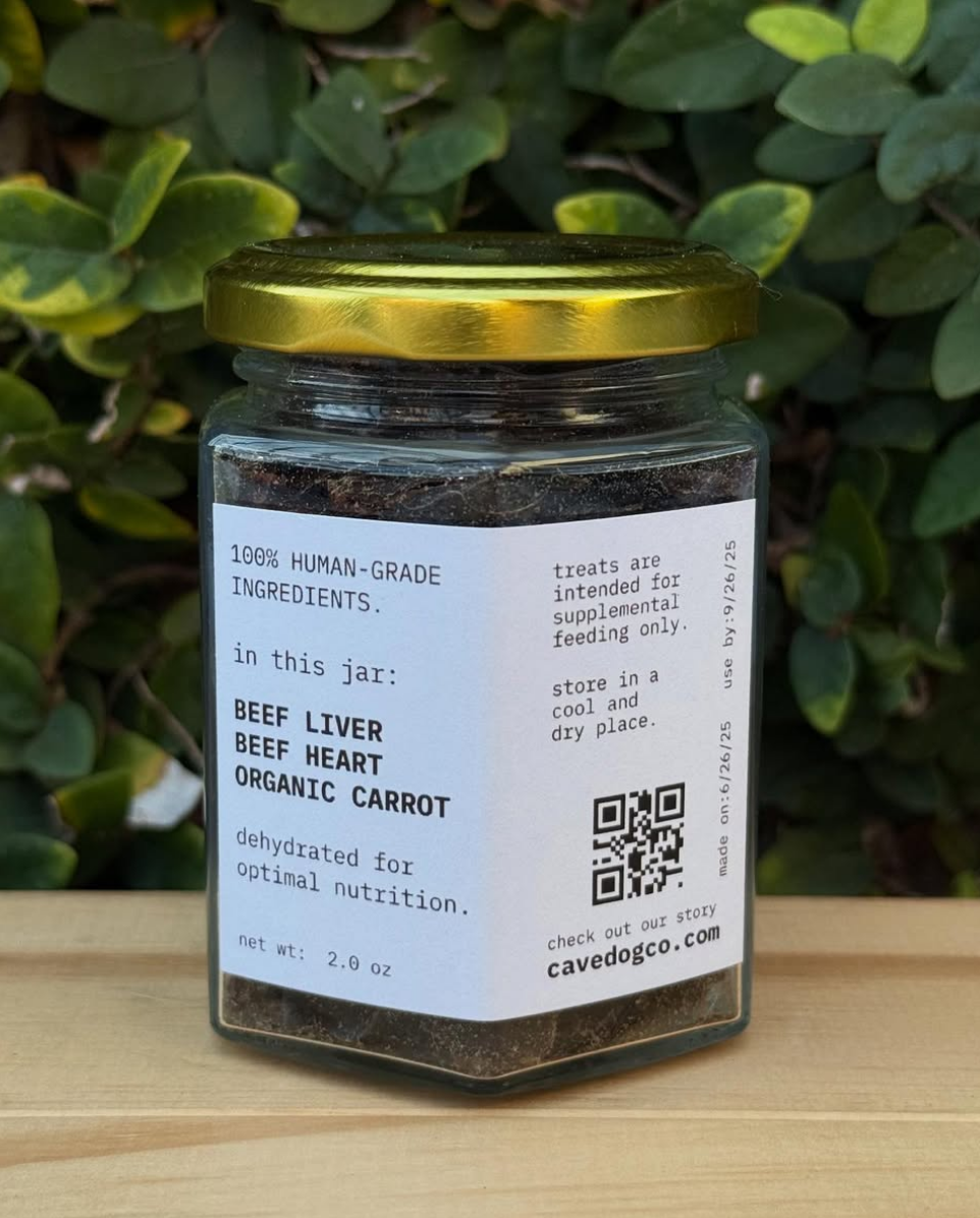 A glass jar of cavedogco.com Beef Liver Beef Heart Organic Carrot dehydrated treats, featuring a gold lid and white label, set on a wooden surface with green foliage in the background.