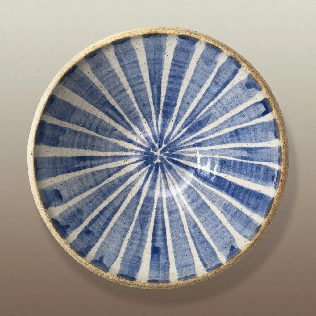 A round ceramic plate with a blue and white radial starburst pattern and a natural beige rim, photographed from directly above on a neutral gray background.