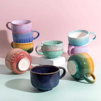 A group of colorful handmade ceramic mugs in various glazes and patterns, arranged on a light blue surface against a soft pink background.
