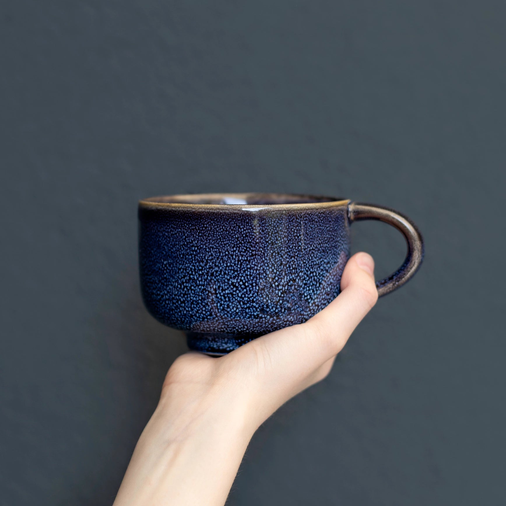 A hand holding a dark blue speckled ceramic cup with a brown rim and handle against a dark gray background.