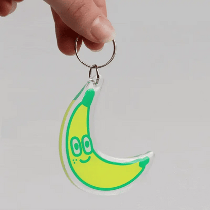 A hand holding a crescent-shaped acrylic keychain charm shaped like a smiling banana, with a silver metal key ring, against a plain light gray background.