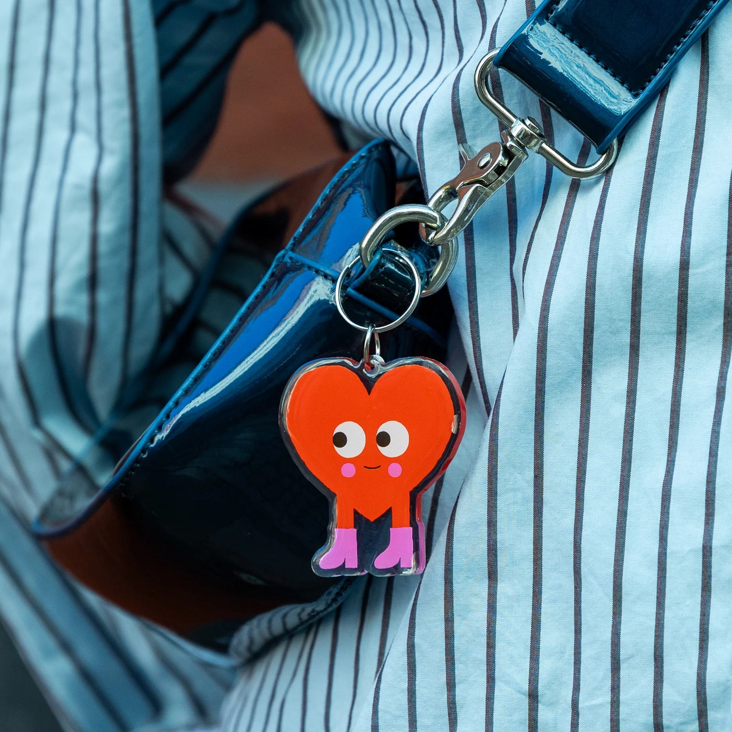 A close-up of a heart-shaped keychain charm with a cute cartoon face and pink legs, attached to a silver clasp and worn over a blue and white striped shirt.