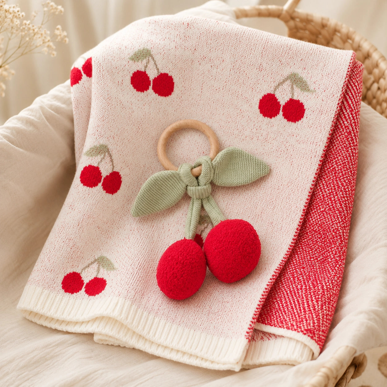 A folded baby blanket or throw in a woven basket, featuring a cream front with red cherry prints, a red textured reverse side, and a wooden ring with a knotted green bow and red pom-pom cherries attached.